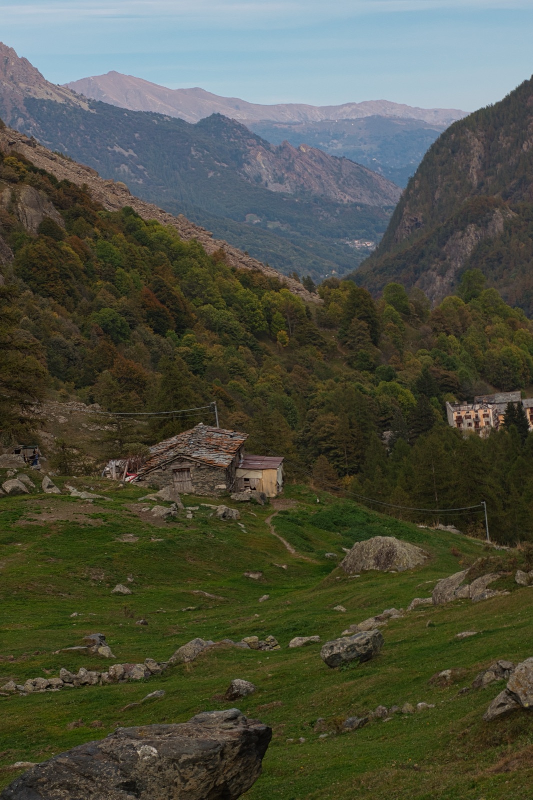 Pascolo con vecchie baite in pietra sul fianco di una valle, con montagne lontane