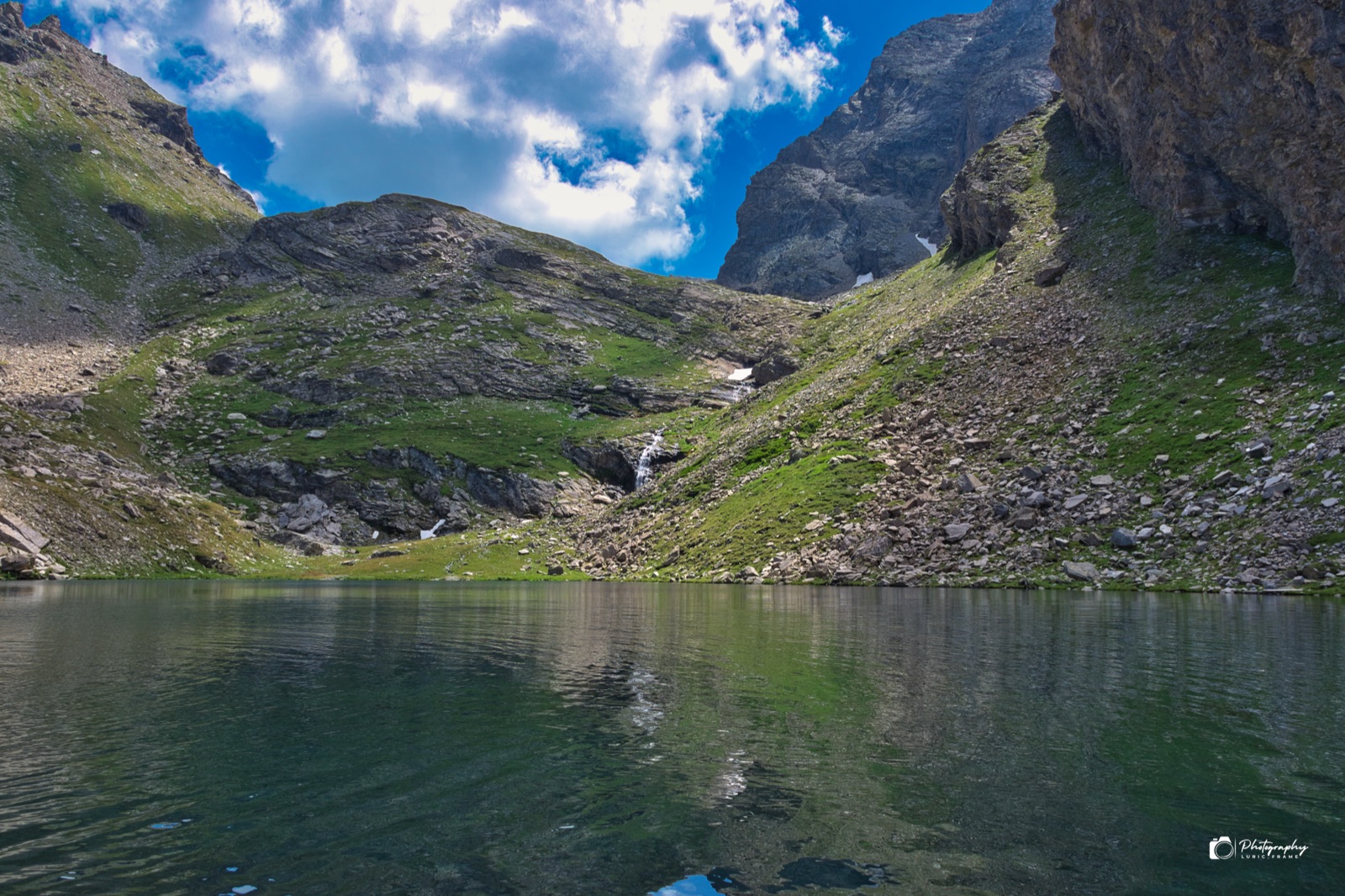 Lago di montagna tra pendii verdi e pareti rocciose, con nuvole bianche nel cielo azzurro