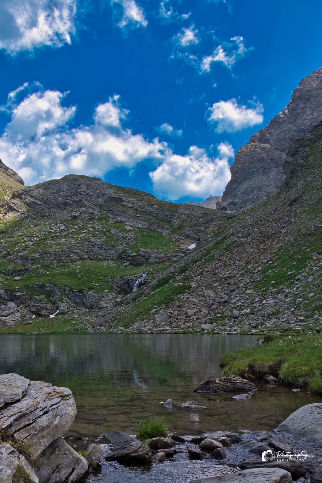Lago alpino con rocce sulla riva, montagne verdi e un cielo azzurro pieno di nuvole