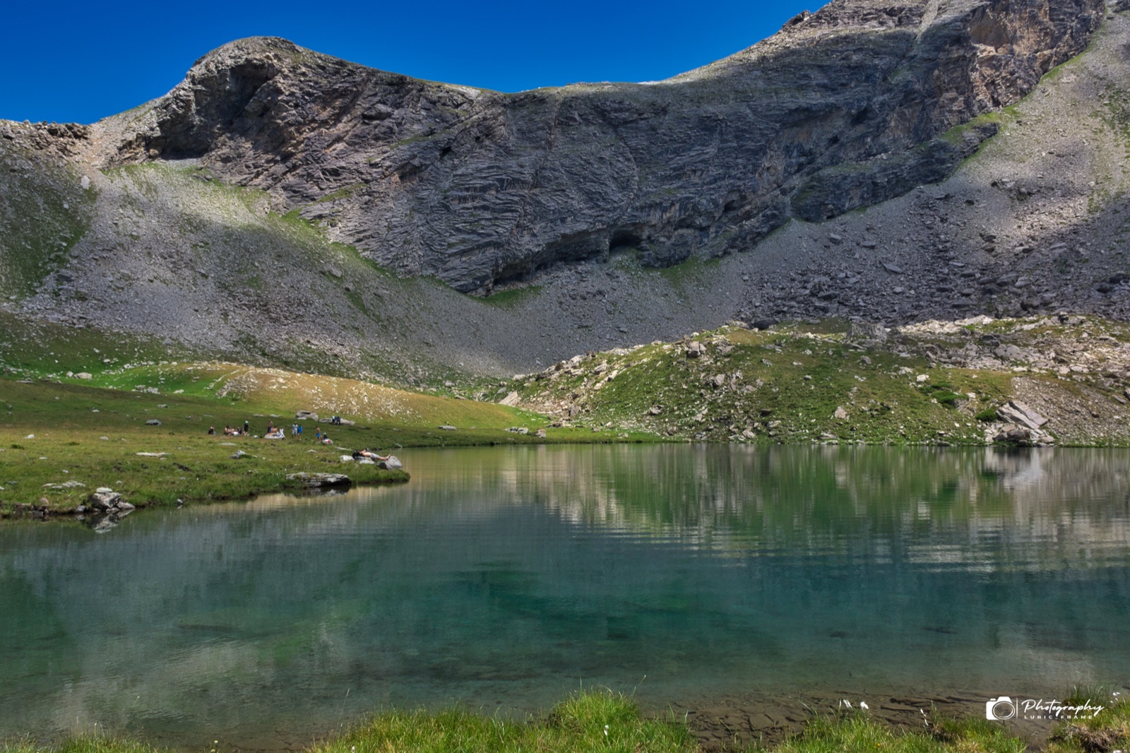 Lago di montagna calmo che riflette una cresta rocciosa e i pendii erbosi