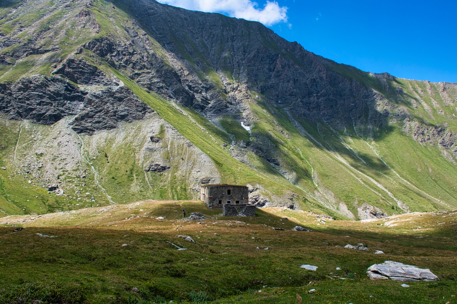 Baita in pietra isolata in un ampio pascolo, sotto una montagna dai pendii erbosi e rocciosi