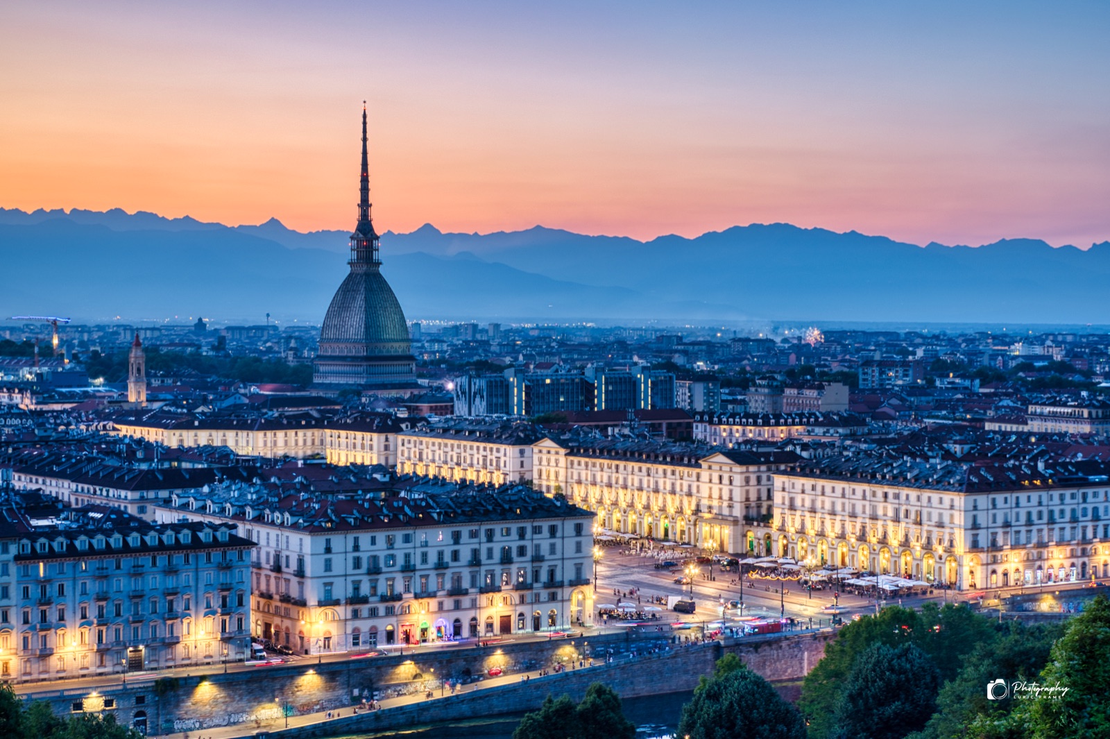 Città all'ora blu vista dall'alto, con un'alta guglia al centro, luci accese e montagne all'orizzonte